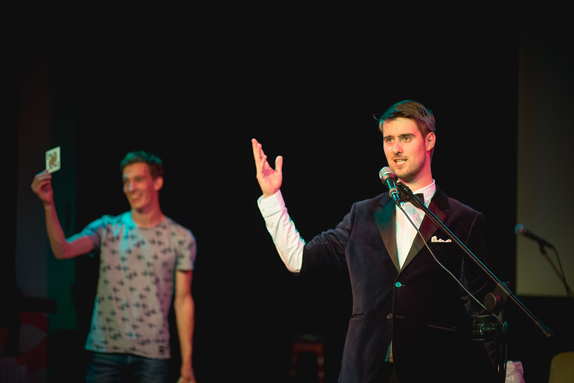 Janse with a suit on a stage talking in a microphone. In the background, a man holds up a playing card. The background of the stage is black.