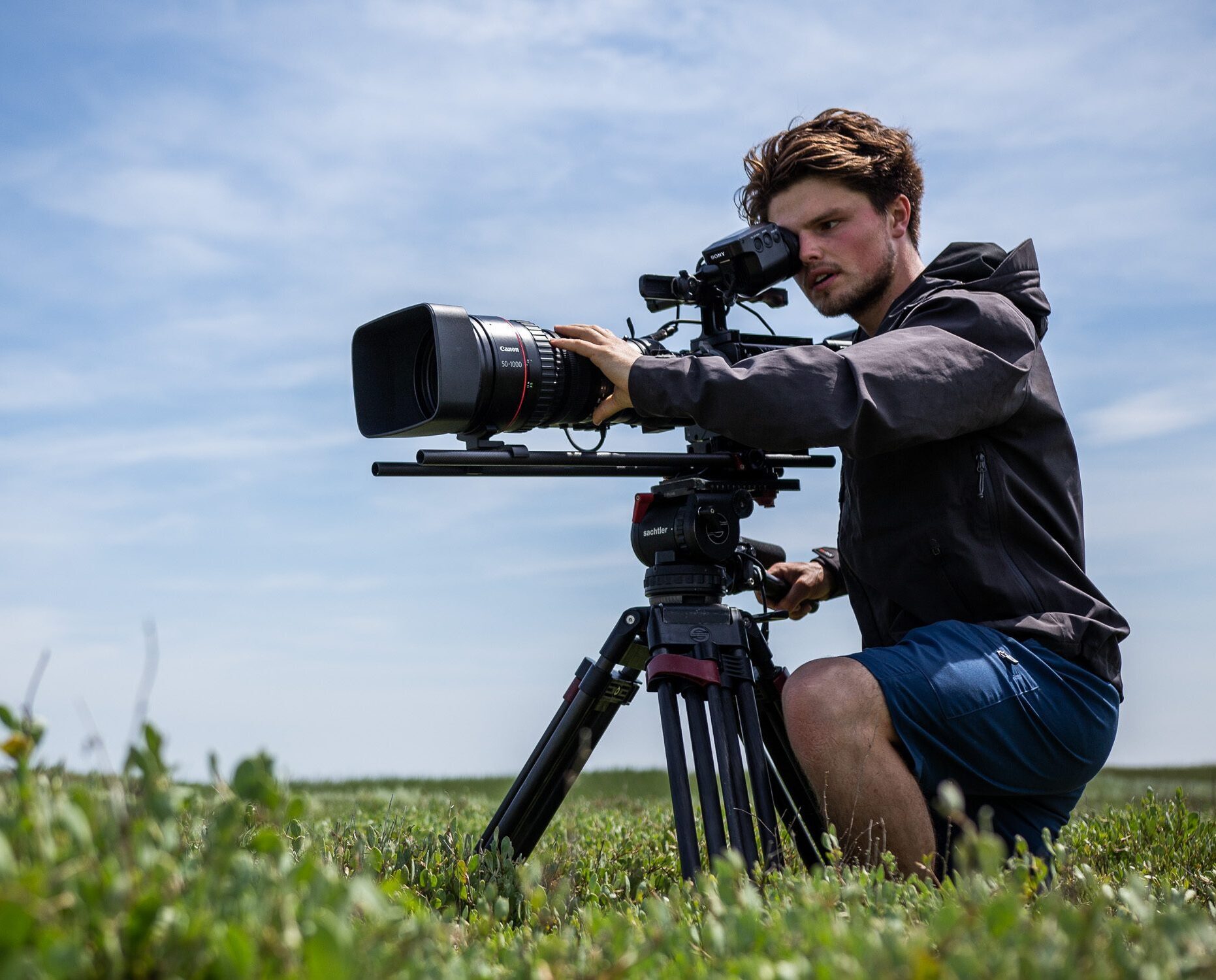 A young man is kneeling in a field. He is looking focused into a big camera that is supported by a tripod. The sky behind him is blue.
