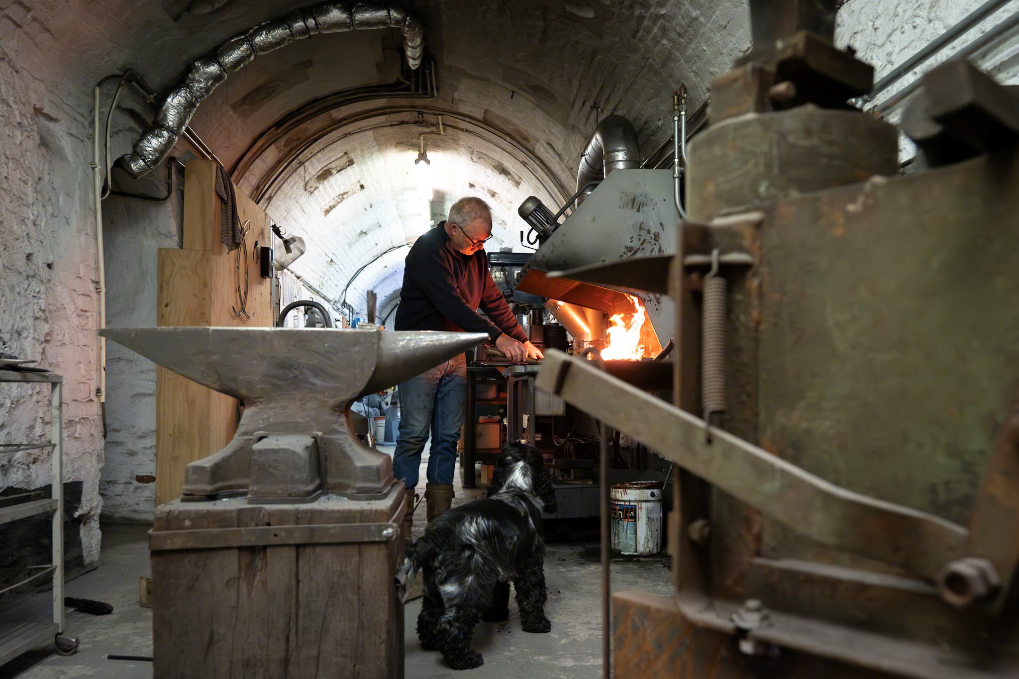 A man working in his smithy, that is located in an arch-shaped cellar. In the foreground, an anvil and machine are placed. In the background, the man working with fire.