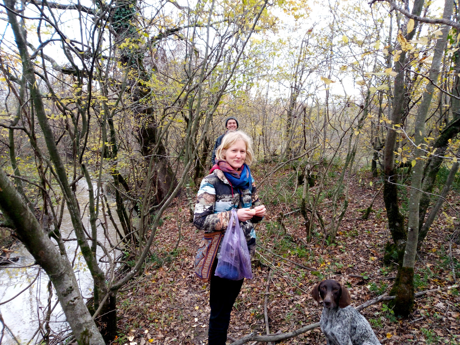 Alumna Anna Samwel in a forest in winter smiling to the camera with a dog on the bottom right and a another person behind her further away also smiling to the camera