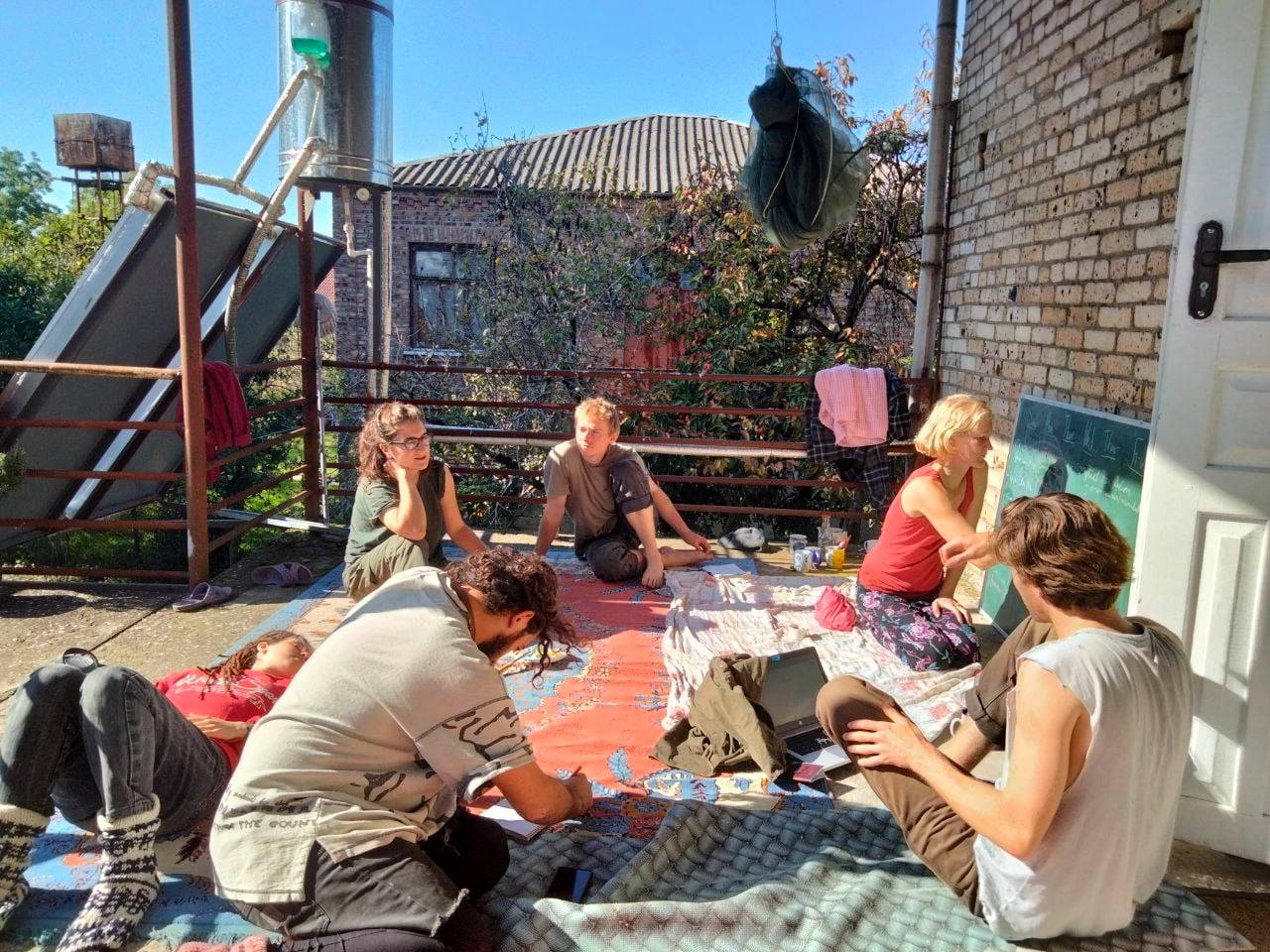 anna samwel siting down on a balcony with 4 other people discussing the ecovillage with volunteers and villagers. she is drawing on a chalkboard while sitting down on carpets. 