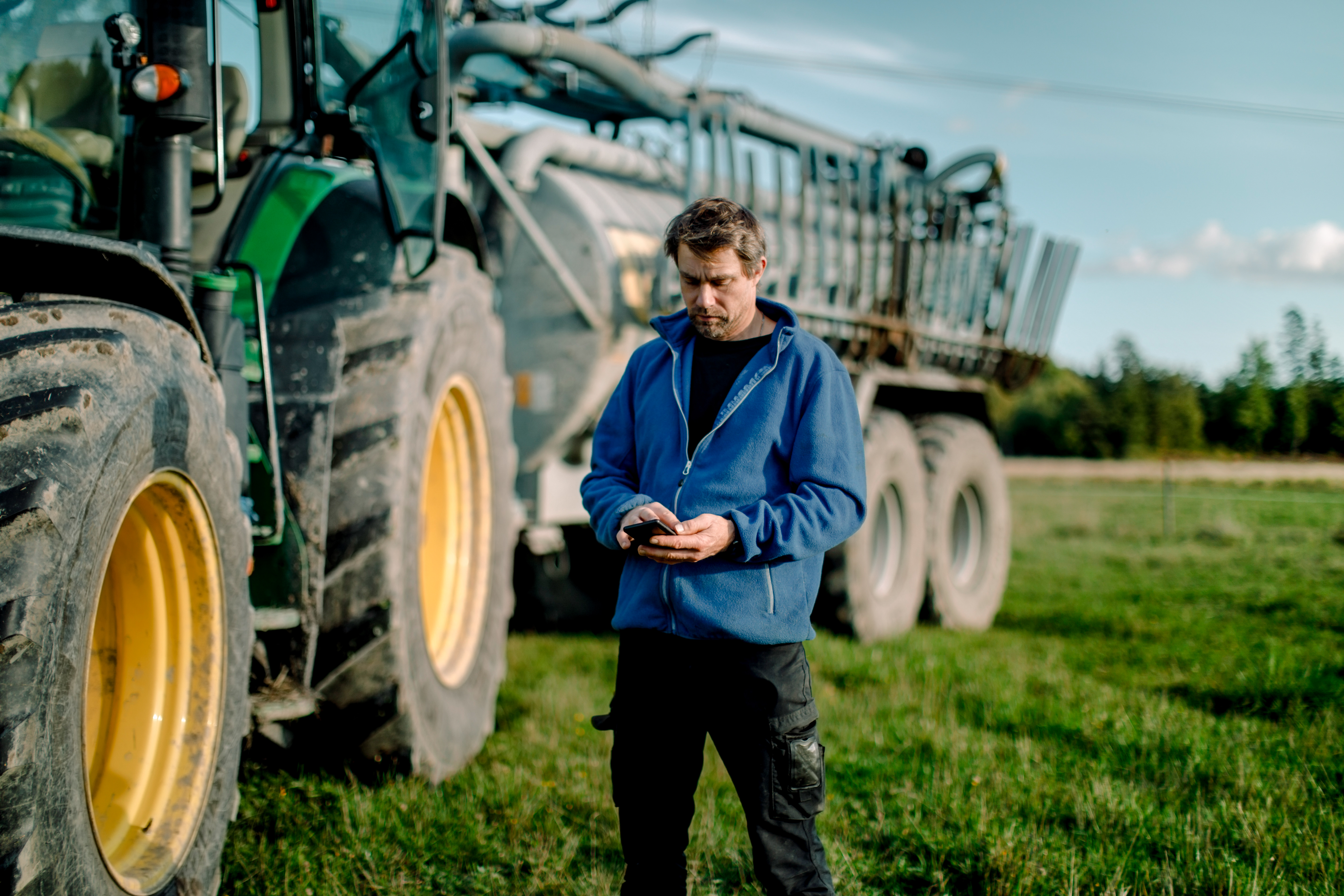 Farmer in a blue jacket looking at his phone. A tractor is in the background. He is standing on a flat farm field