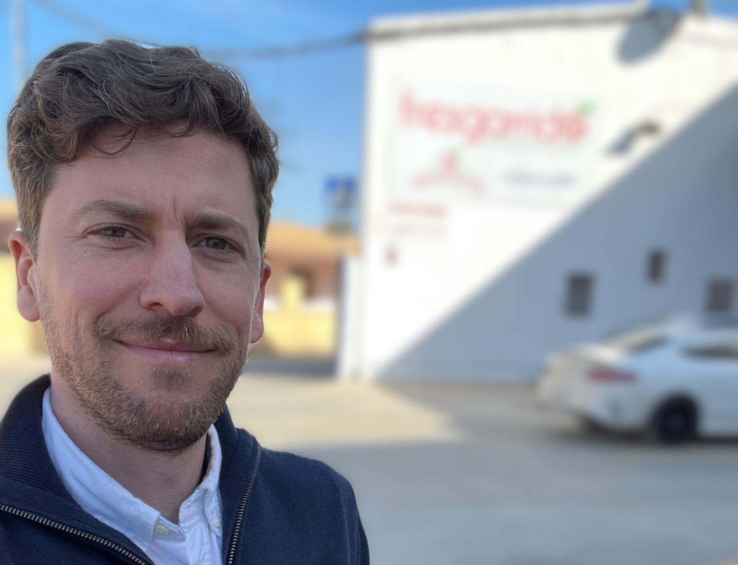A young men looking towards the camera. In the background a big building and a car