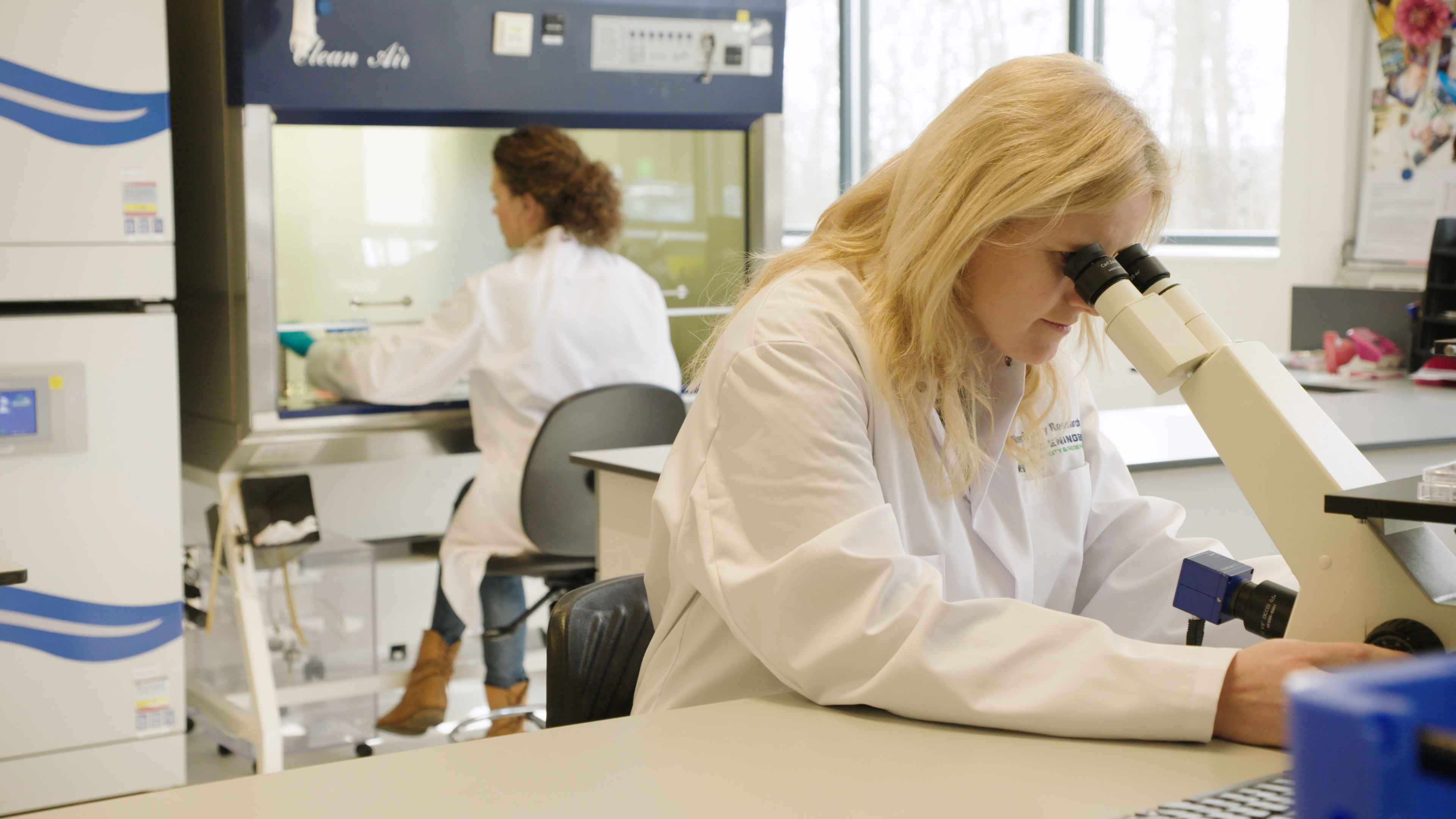 Two female researchers are working in the lab. A young blonde woman is looking through a microscope in the foreground, while another woman is working under a fume hood in the background.