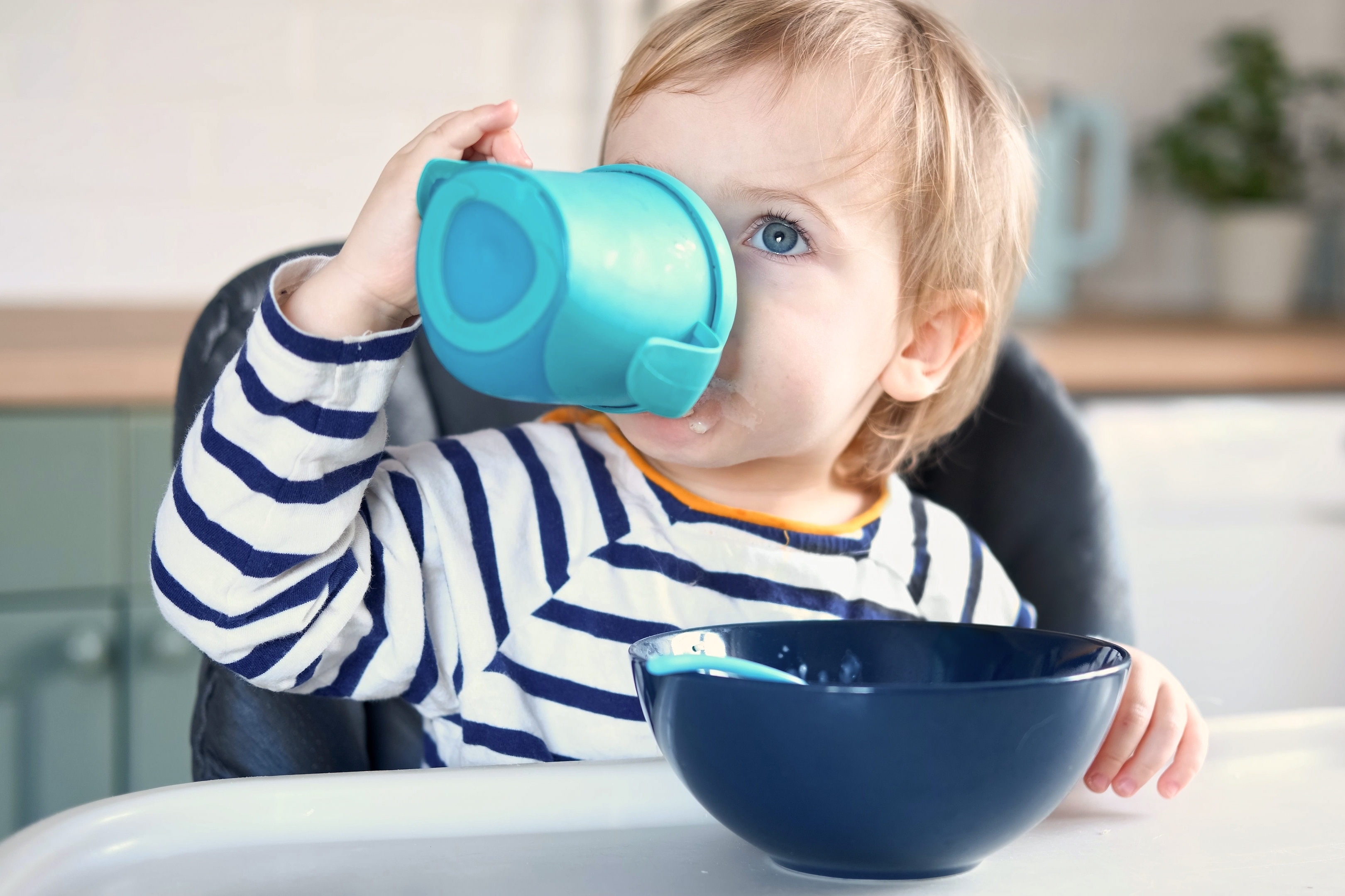 Een peuter gekleed in een gestreept shirt, zit in een kinderstoel aan tafel en drinkt uit een flesje.