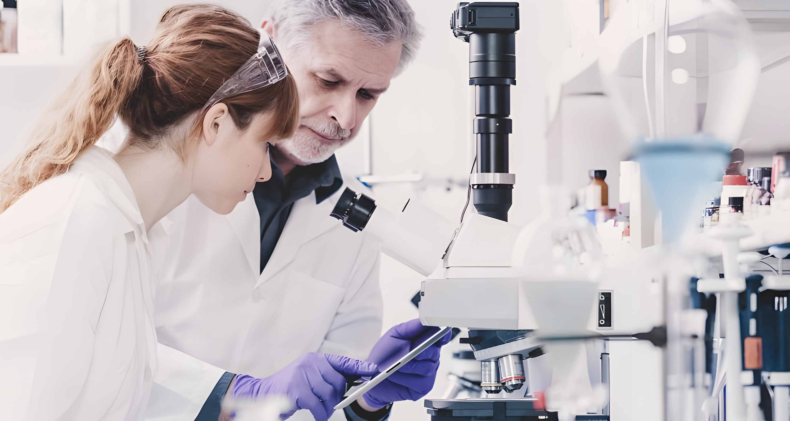 Two researchers stand next to a microscope and look at the screen of a tablet