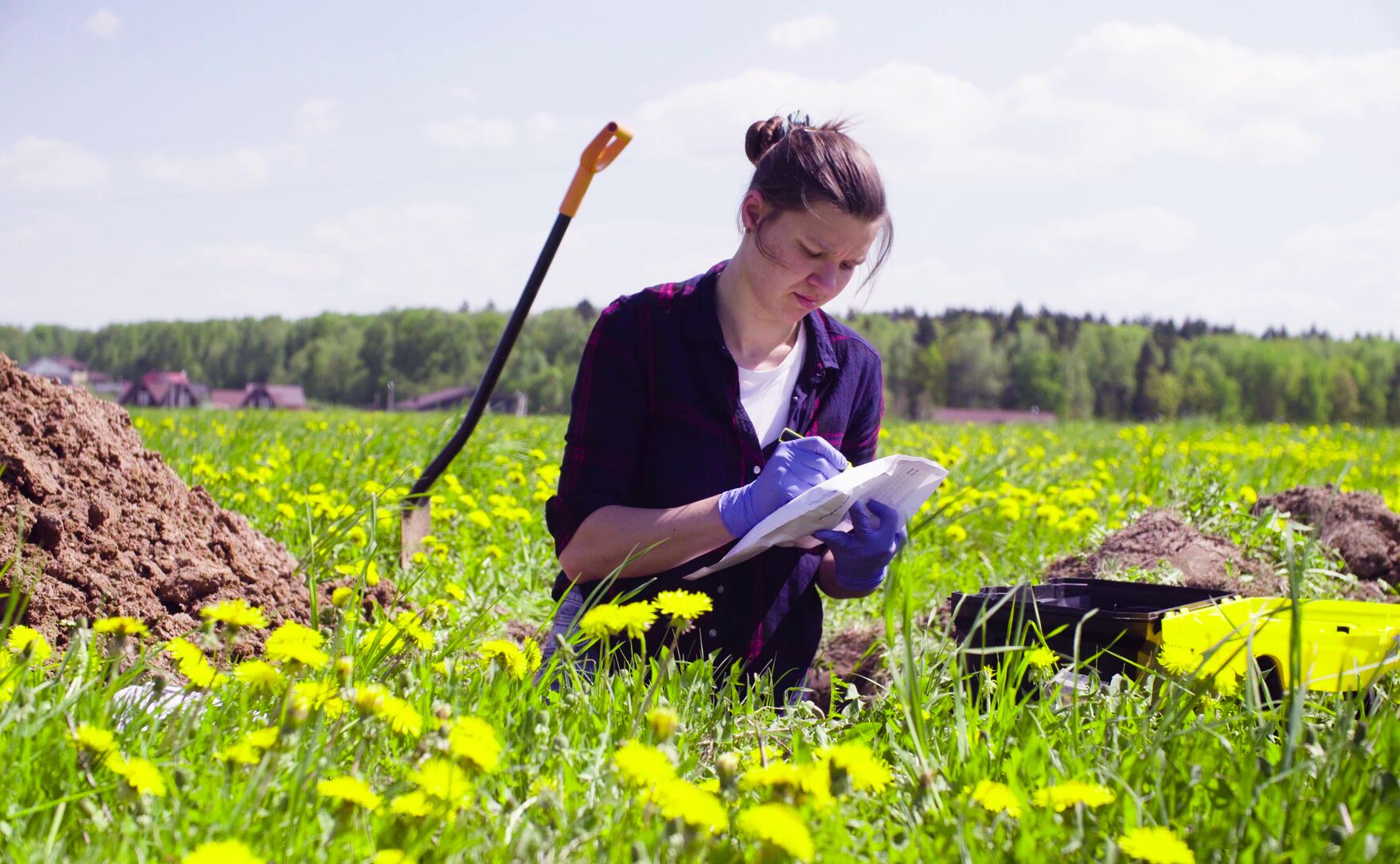 A woman making notes, while sitting in a field filled with flowers and a shovel in the background