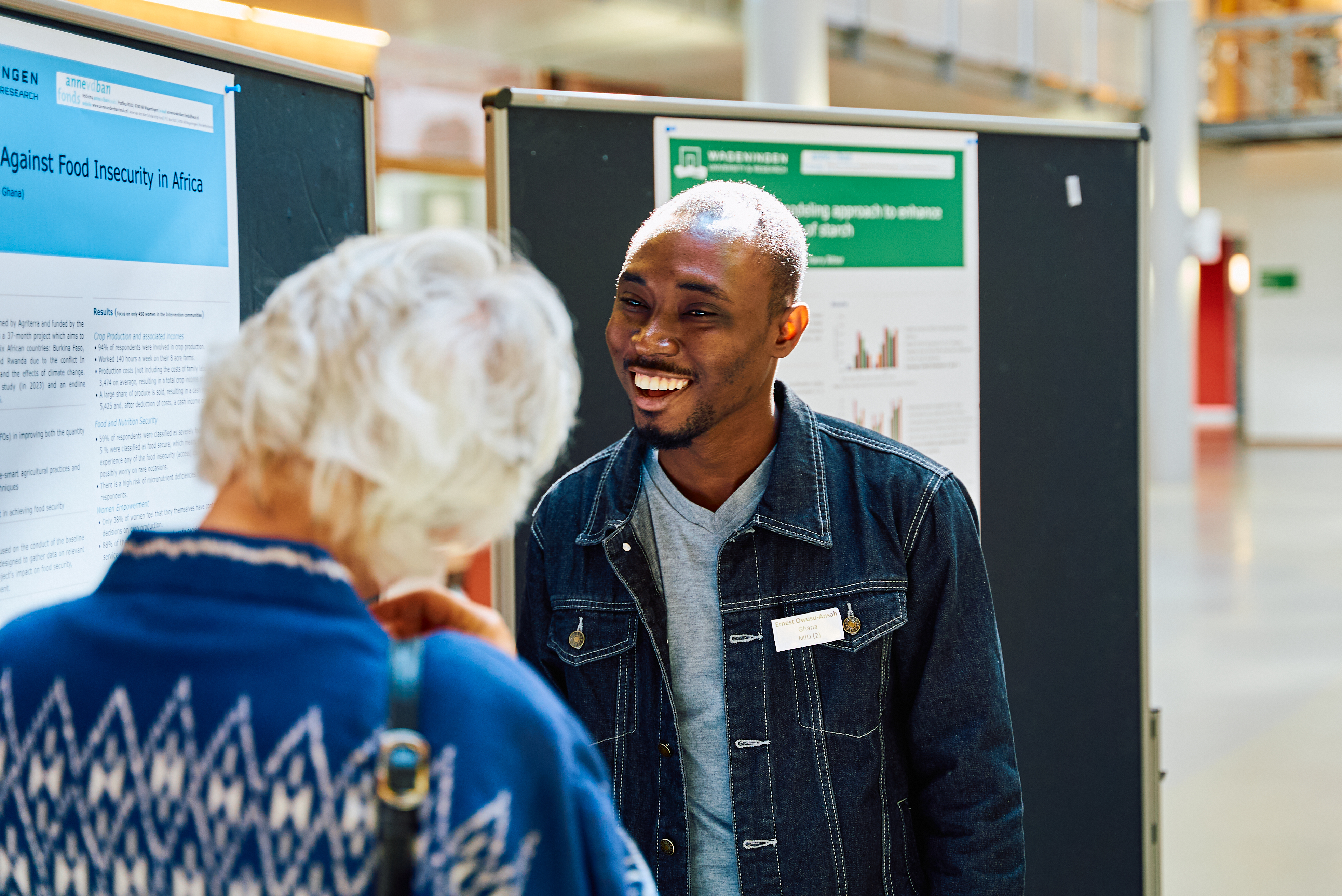 Een student lacht samen met een oudere vrouw, die van achteren te zien is. Op de achtergrond hangen wetenschappelijke posters.