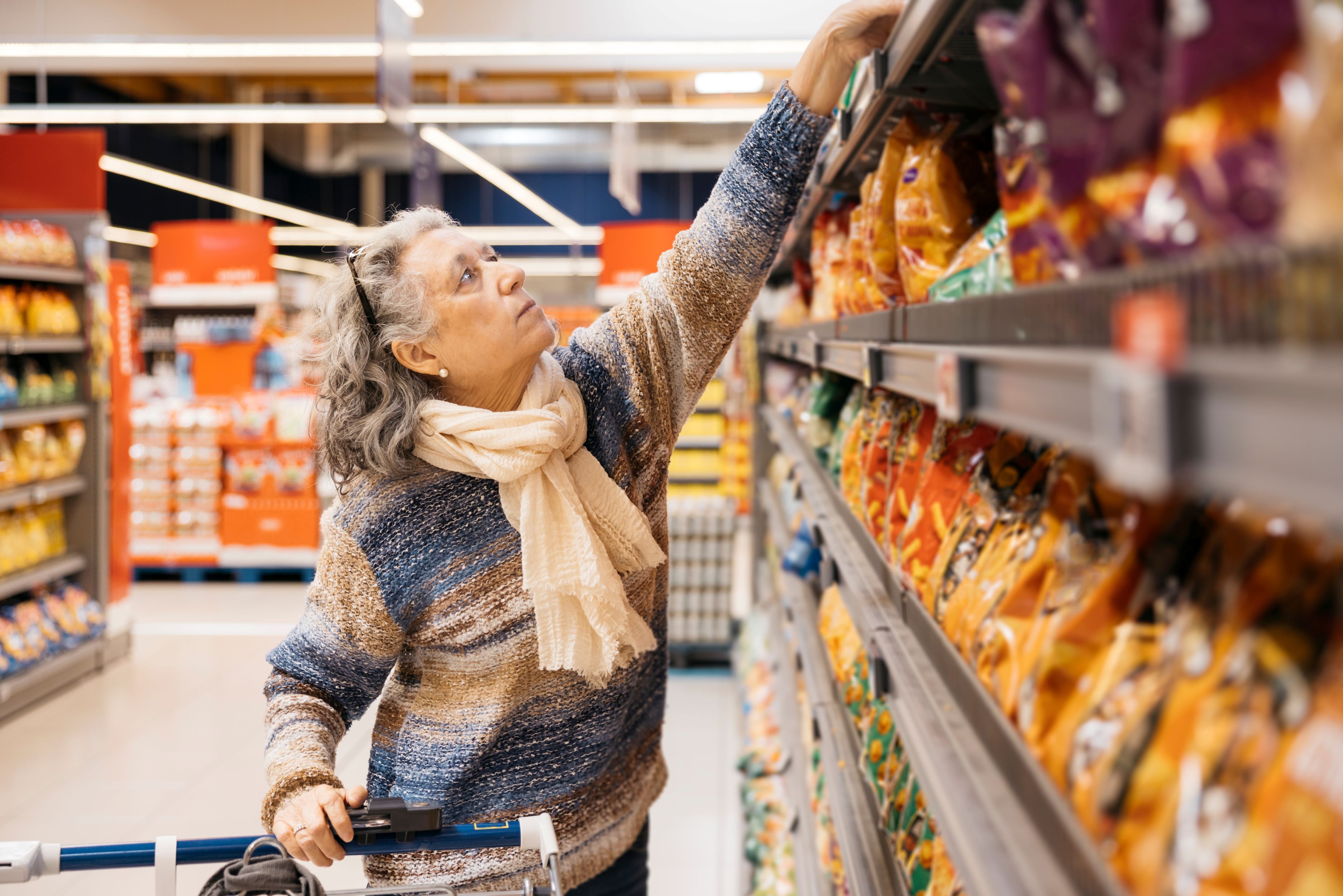 Senior woman shopping for groceries. She picks a snack food from a high shelf in a store
