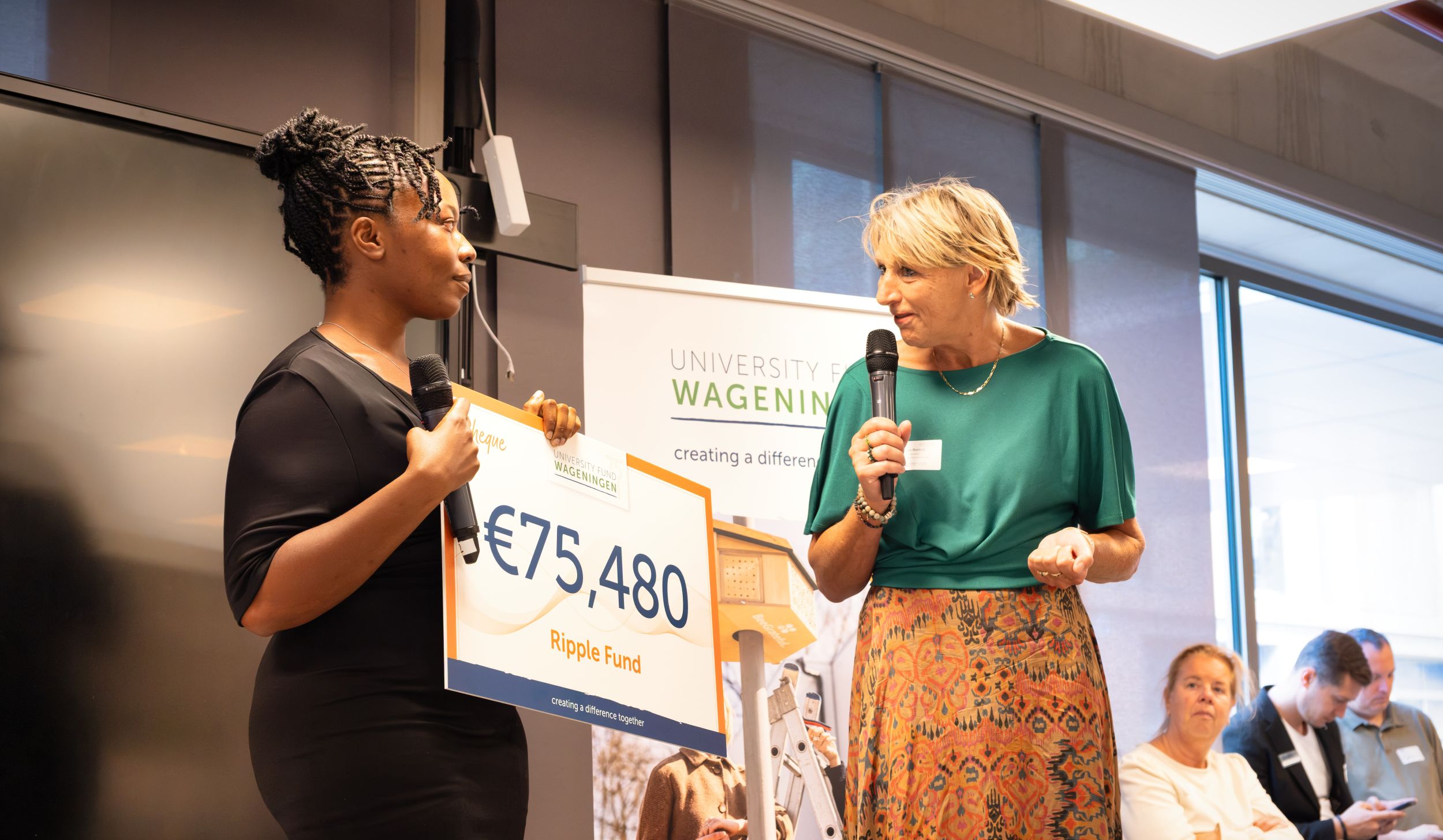 Twee mensen op een podium, een houdt een cheque vast van €75.480 en de andere een microfoon