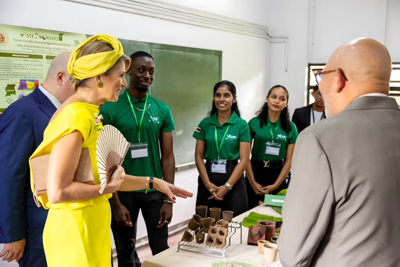 Queen Maxima, accompanied by two men in suits, is looking at plant pots on a table in a classroom. Three young people wearing the same green T-shirts are standing by the plant pots.
