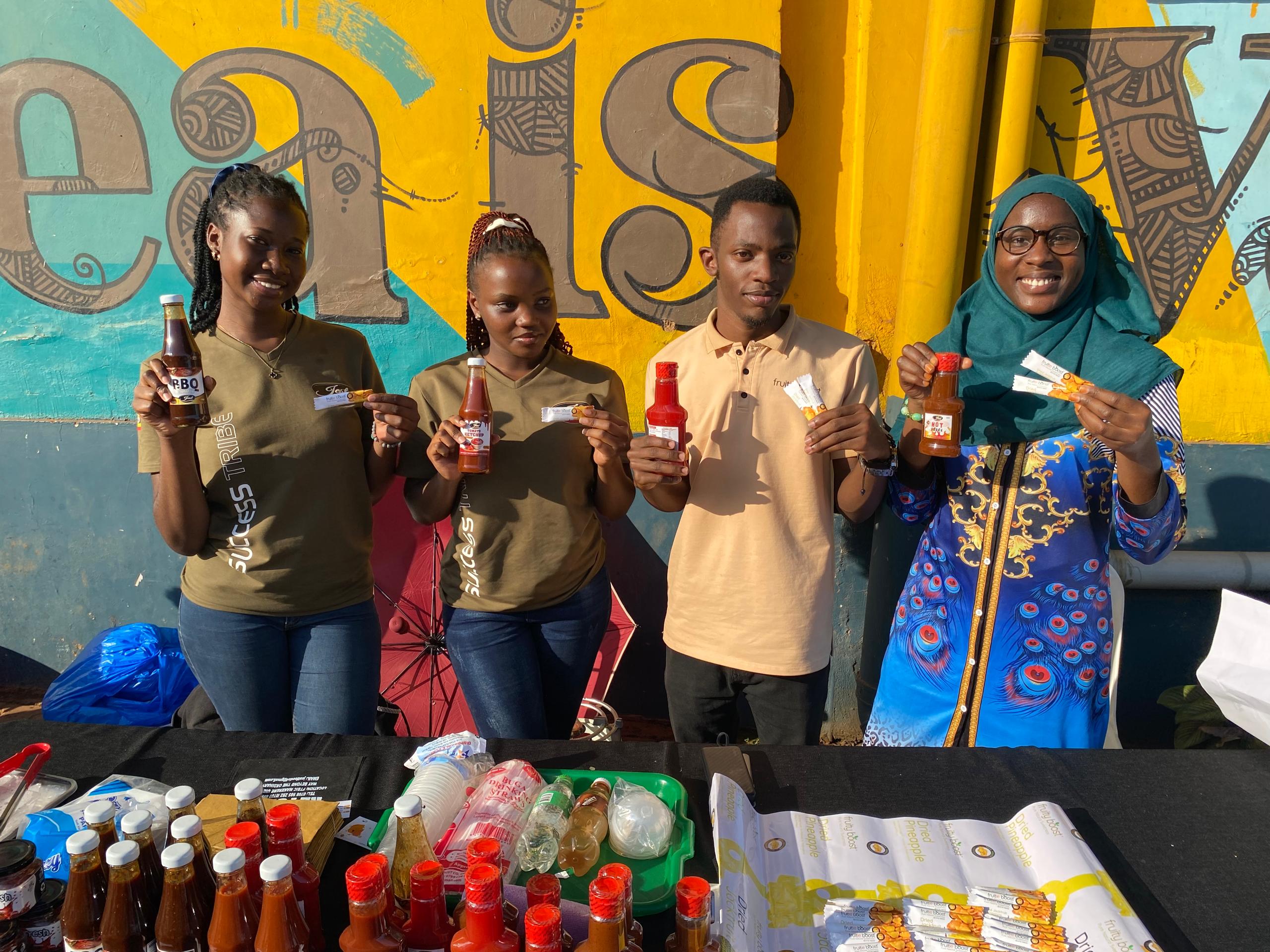 Four african young people presenting a sauce bottle behind a stand, in front of a colourful wall