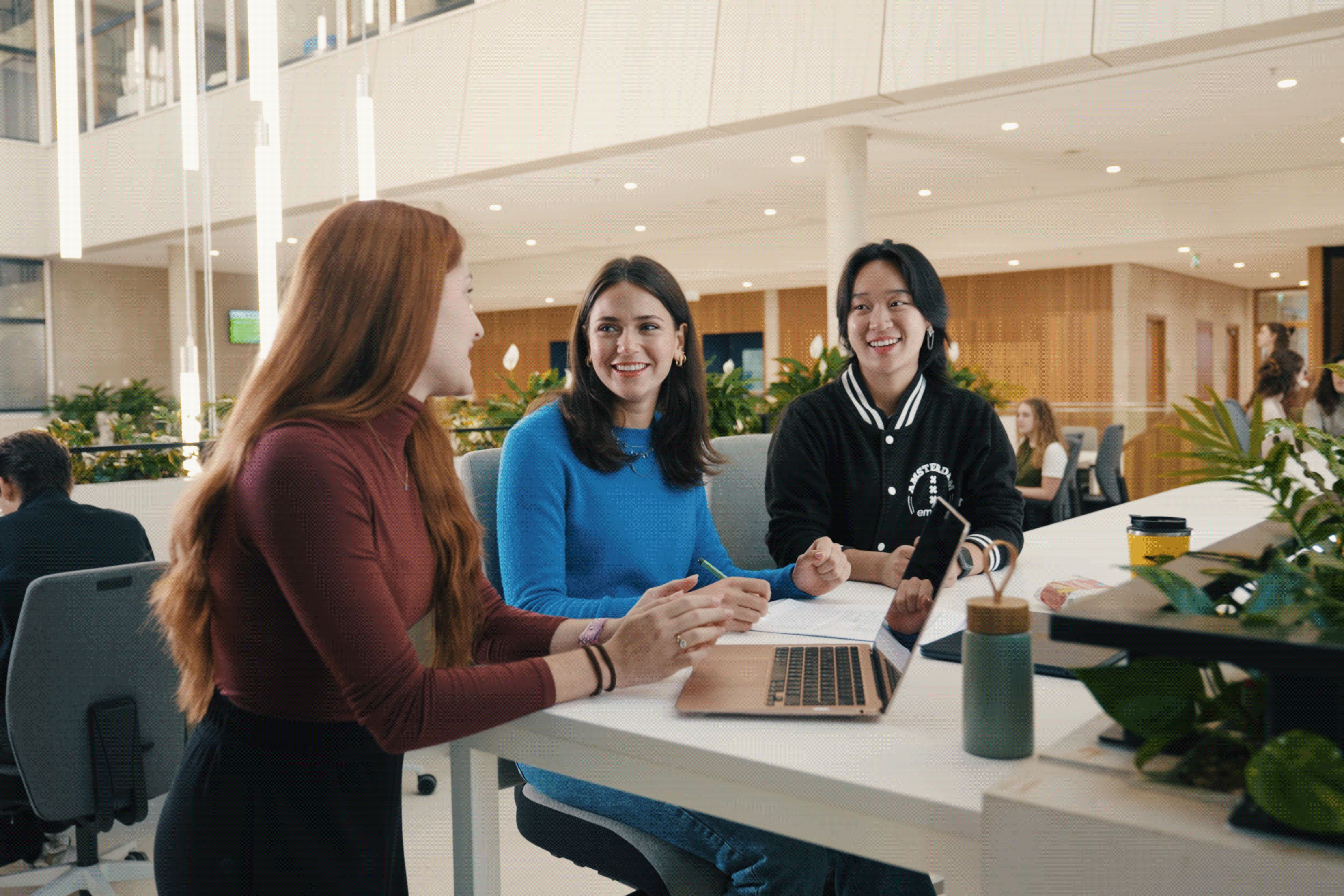 Drie studenten zitten samen aan een tafel met een laptop en praten met elkaar in een lichte, moderne studieruimte op de campus.