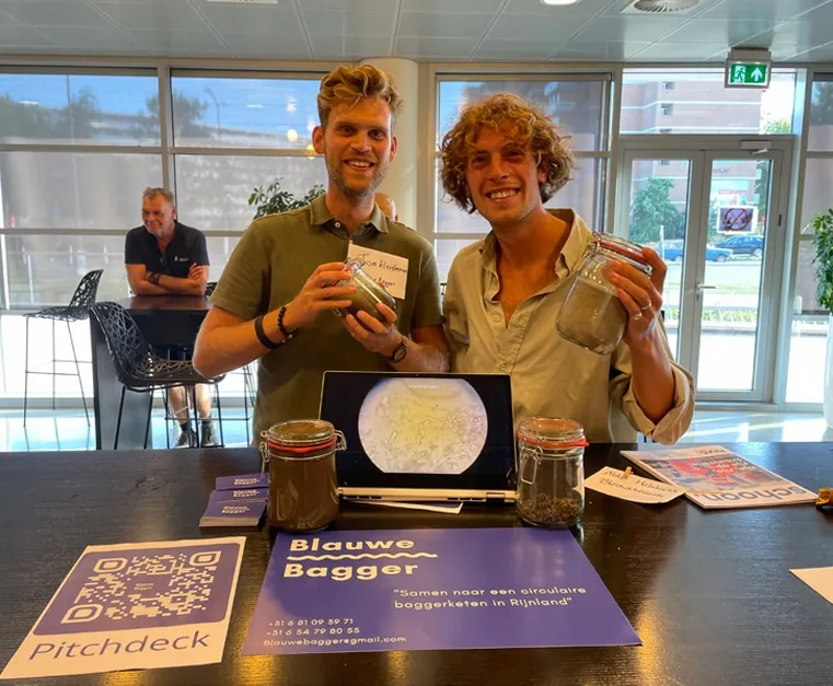 Tom and his co-worker hold a jar with dredge in front of a "Blauwe Bagger" sign