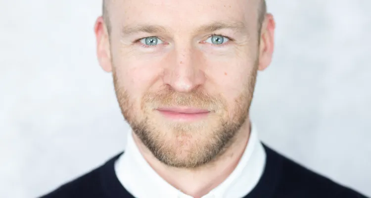 Close up picture of Cedric smiling for the camera. He has a light-brown beard and blue eyes, he is wearing a black sweater with a white collar.